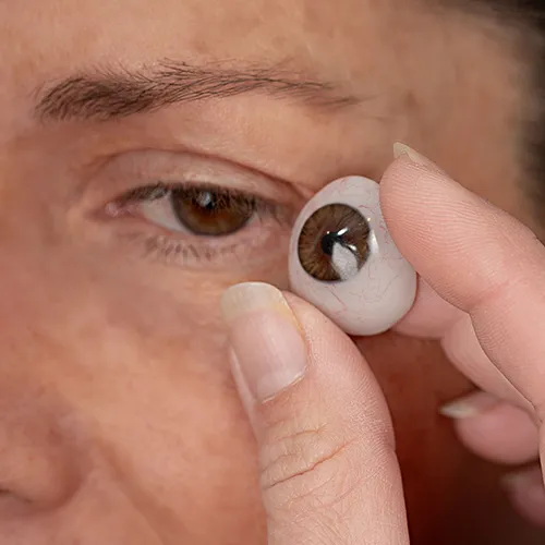 Pessoa segura uma prótese ocular marrom próxima ao olho de uma segunda pessoa, em movimento de colocação. A imagem destaca o detalhe do dispositivo e o processo de adaptação estética.