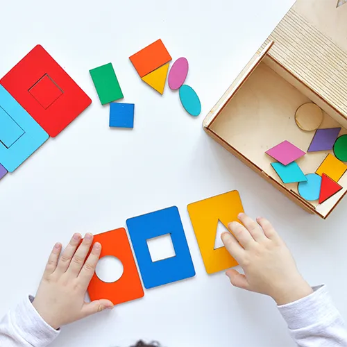 Mãos infantis manipulam peças coloridas de encaixe em diferentes formas geométricas, sobre uma mesa com caixa de madeira aberta ao lado. A cena ilustra uma atividade de estimulação visual, que contribui para o desenvolvimento cognitivo e motor da criança.