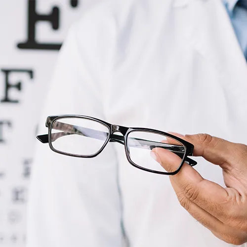 Óculos com prisma sendo exibidos por profissional da saúde, com tabela oftalmológica desfocada ao fundo.