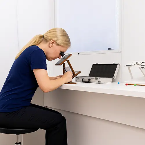 Mulher de cabelo loiro preso realiza exercícios de ortóptica sentada em uma mesa, utilizando equipamentos específicos para treinamento visual. A cena representa práticas voltadas à reabilitação e ao fortalecimento da coordenação ocular.