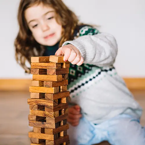 Criança de cabelos castanhos e cacheados brinca com blocos de madeira empilhados, exercitando coordenação motora fina e concentração. A atividade pode estar associada a práticas de fisioterapia ocular, que estimulam a visão, o foco e a percepção espacial durante o desenvolvimento infantil.